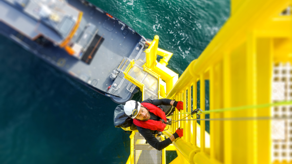 A male offshore worker wearing a high vis and hard hat, climbing up a ladder on a ship.