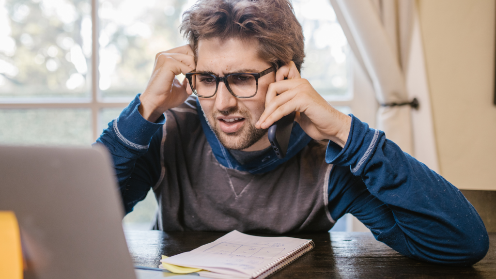 A man sat at his work desk at home, with a notebook and his laptop in front of him. He's also on his mobile phone looking confused.