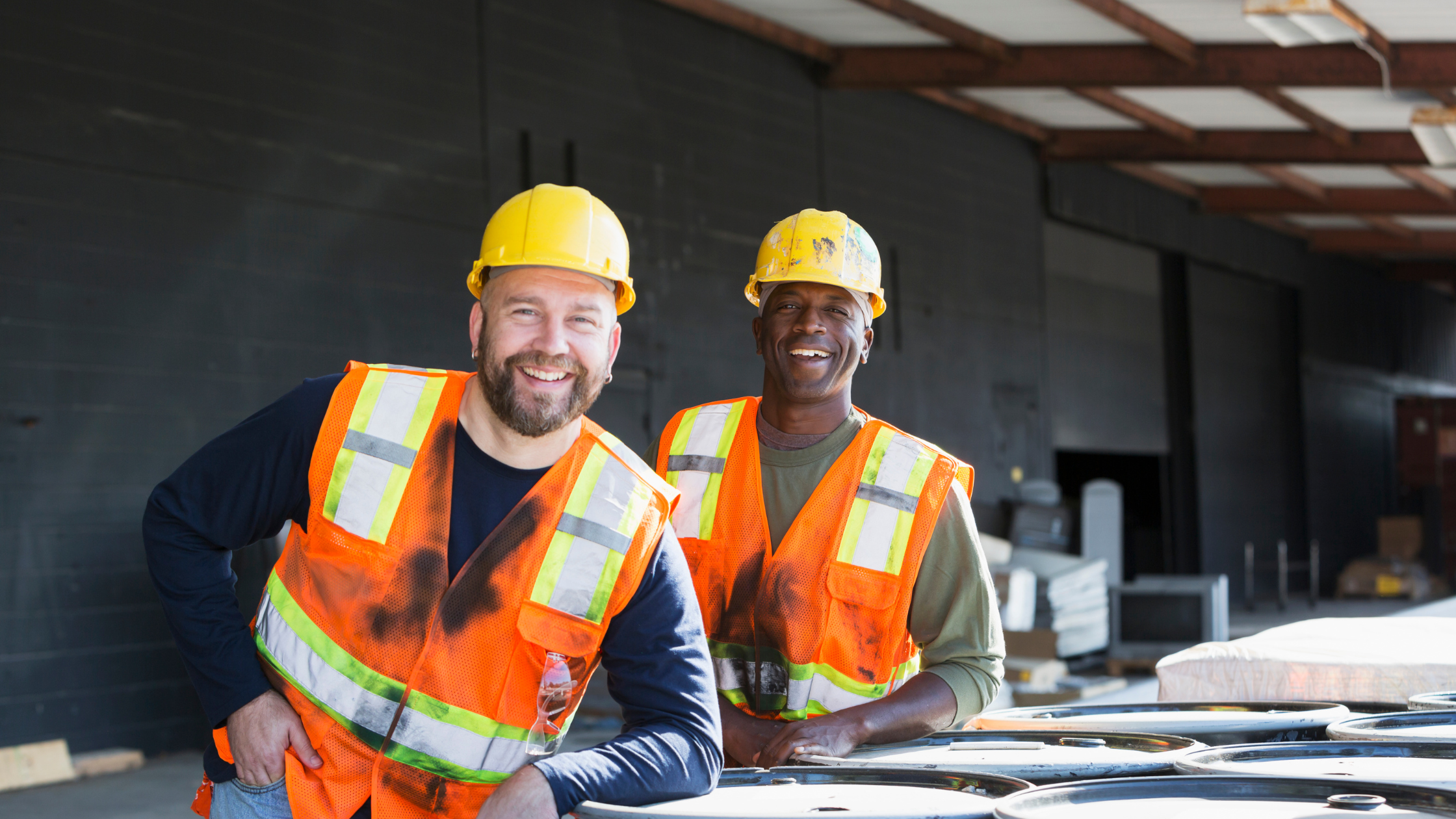 Two construction workers in high vis vests and yellow hard hats smiling at the camera.